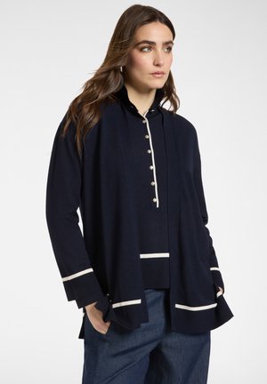 Femme portant un haut marine à manches longues avec des boutons en perle et une bordure blanche, associée à un jean bleu, les mains dans les poches, regardant sur le côté.