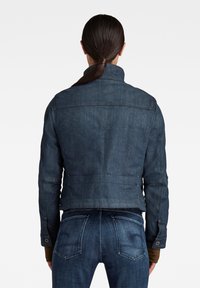 Tmavě modrá džínová bunda s límcem, knoflíkovými detaily a přiléhavým střihem. Viditelné švy a kratší délka vzadu.