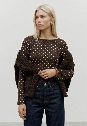 Femme aux longs cheveux blonds portant un haut à manches longues marron à pois, un jean sombre et un pull sombre drapé sur ses épaules.