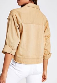 Lyhennetty beige farkkutakki, jossa on rullatut hihansuut, klassinen kaulus sekä ompeluyksityiskohtia harteilla ja helmoissa.