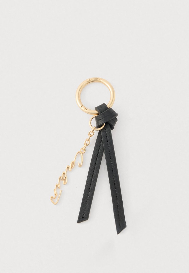 Porte-clés Calvin Klein doré avec boucle en cuir noir et pendentif en script doré suspendu à une chaîne sur fond blanc.