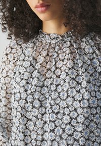 Femme aux cheveux bouclés portant une blouse transparente à motifs floraux avec de petites fleurs blanches et bleu clair et un col montant volanté.
