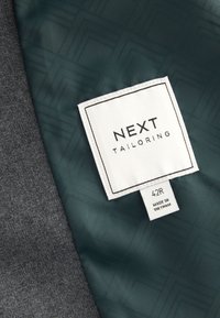 Étiquette de couture indiquant "NEXT TAILORING" et taille "42R" sur un tissu vert foncé à motif à côté d’un tissu en laine gris. Fabriqué au Vietnam.