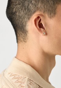 Zlaté náušnice s krištáľovou kamennou ozdobou, nosené v uchu, na pozadí krátkych, štruktúrovaných vlasov a svetlosivého pleteného goliera.