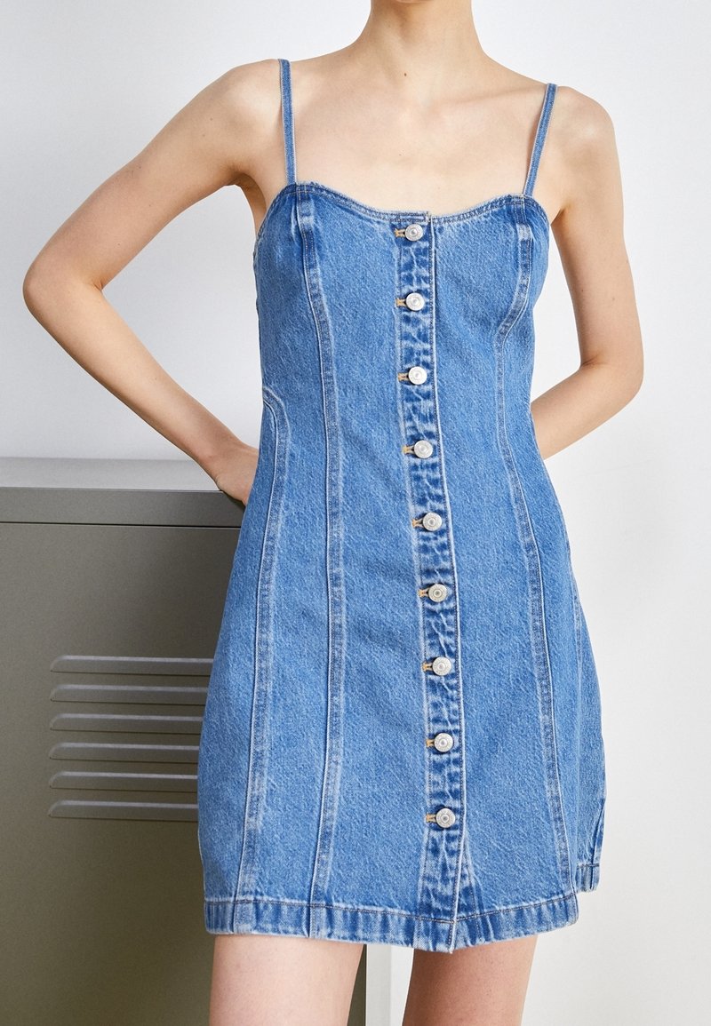 Femme portant une robe en jean bleu sans manches avec des boutons sur le devant, debout avec les mains derrière le dos sur un fond clair.