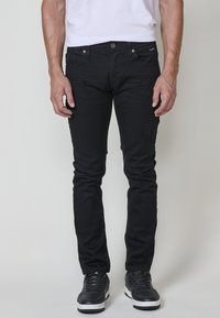 Crne slim-fit traperice s glatkom površinom, pet džepova i suptilnim istrošenjem. Uparene s crnim sportskim cipelama i običnom bijelom majicom.