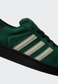 Zielony zamszowy sneaker z czarną podeszwą i sznurówkami, z trzema białymi paskami i słowem "SUPERSTAR" wytłoczonym na boku.