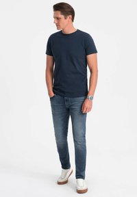 Tmavě modré bavlněné tričko s kulatým výstřihem, spárované se světle modrými džínami. Model nosí bílé tenisky s hnědými akcenty.