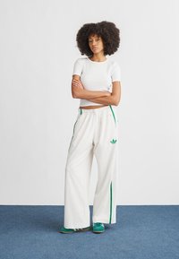 T-shirt branca cropped, calças de fato de treino brancas largas com riscas verdes nas laterais e logótipo. Modelo usa ténis verdes. Fundo liso, chão azul.