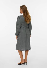 Mujer con cabello largo vistiendo un vestido midi a cuadros en blanco y negro y sandalias de tacón negras, de pie de perfil izquierdo sobre un fondo blanco.