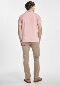 Jasnoróżowa koszulka polo z białymi wykończeniami, noszona z beżowymi spodniami i casualowymi brązowymi butami, o prostym wzorze i miękkiej fakturze materiału.
