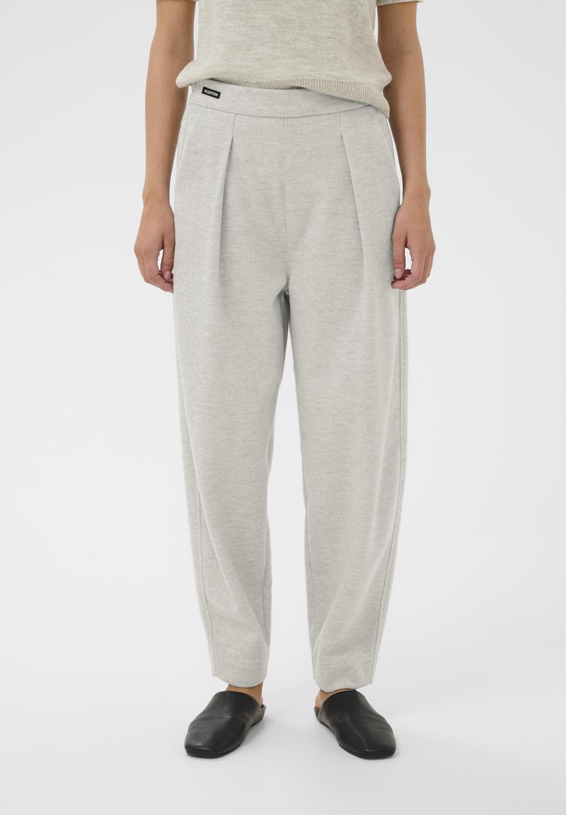 Világosszürke, plisszírozott nadrág, puha anyagból készült. Széles derékpántja és beszűkült lábai vannak. Egyszerű fekete slip-on cipővel párosítva.