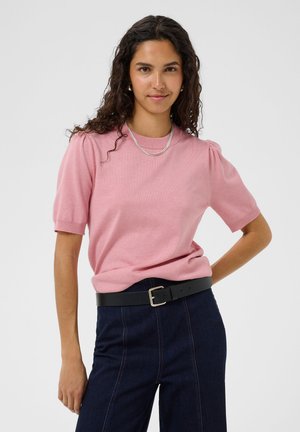 Jeune femme aux cheveux bouclés portant un haut rose à manches courtes, un pantalon foncé taille haute et une ceinture noire, debout avec une main sur la hanche.