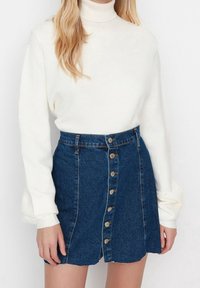 Vrouw in een witte coltrui en een hoog-waisted blauwe denim rok met knoopsluitingen aan de voorkant.