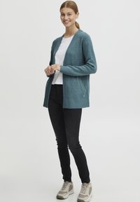 Turkos stickad kofta med en texturerad yta, öppen fram, ribbad nederkant och två sidofickor, kombinerad med svarta jeans och ljusa sneakers.