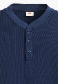 Marineblaues, geripptes Henley-Shirt mit rundem Kragen, dreiknöpfiger Blende und strukturierter Stoff. Etikett am Halsausschnitt sichtbar.
