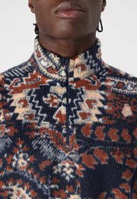 Pile con cerniera in blu navy, crema e ruggine con motivo geometrico. Tessuto morbido con collo alto e dettaglio con logo ricamato.