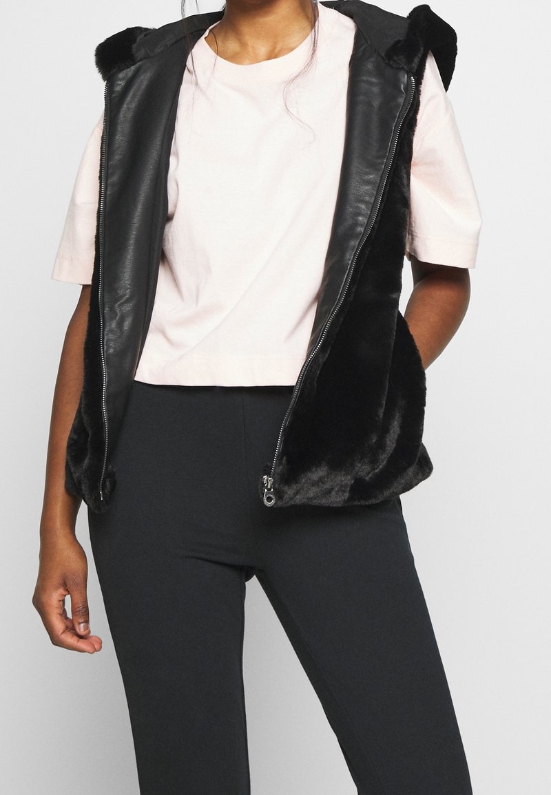 Black leather vest with faux fur trim, featuring a zip closure, two side pockets, and a hood. Worn over a light pink cropped t-shirt.