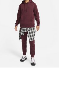 Burgundy hoodie with front pocket, black logo pattern; plaid shirt tied around waist, matching pants, and black/white sneakers.