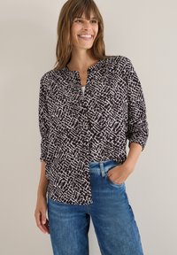 Sort-hvid mønstrede bluse med krave, knaplukning foran og oprullede ærmer, parret med blå denimjeans.