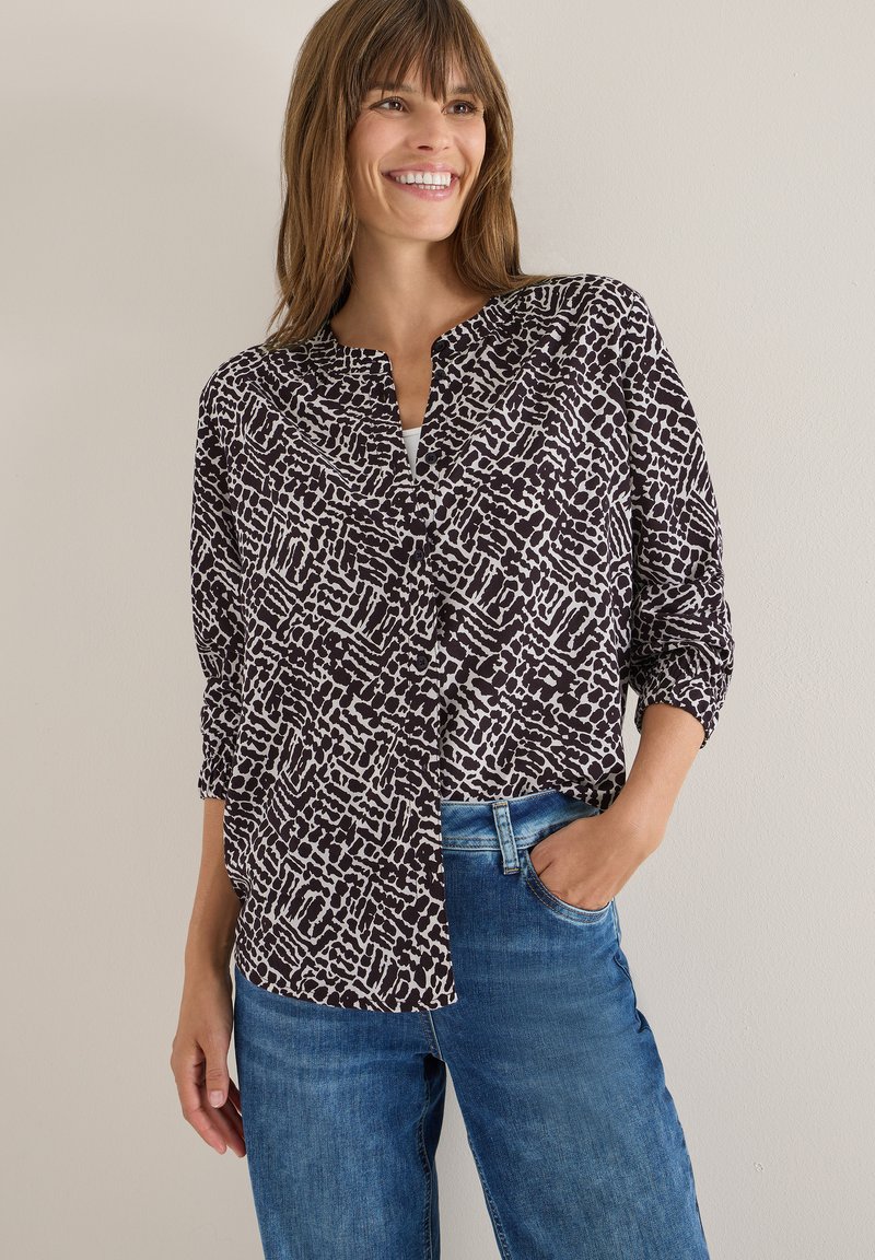 Sort-hvid mønstrede bluse med krave, knaplukning foran og oprullede ærmer, parret med blå denimjeans.