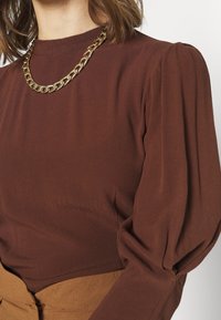 Haut marron à manches longues avec un col montant et des épaules bouffantes, fabriqué dans un tissu doux. Associé à un collier chaîne en or massif.