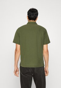 Camicetta verde oliva a maniche corte con colletto, presenta un orlo dritto e una vestibilità rilassata. Realizzata in tessuto leggero con una texture liscia.