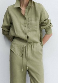 Oliwkowa, lniana koszula zapinana na guziki z kieszonką na piersi i podwiniętymi rękawami, w zestawie z pasującymi spodniami z elastycznym paskiem. Gładka faktura.