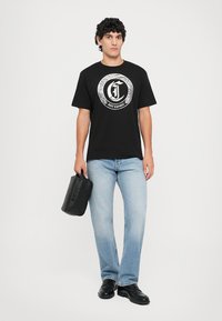 Hombre vistiendo una camiseta con logo Just Cavalli negra, jeans claros, zapatos negros, sosteniendo un estuche de cuero negro con la mano derecha, de pie sobre un fondo blanco.