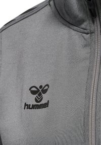 Grå zip-up jakke lavet af tekstureret stof, med et sort Hummel-logo centralt placeret på brystområdet langs lynlåsen.