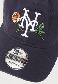 Tumši zils cepure no faktūras auduma ar baltu izšūtu "NY" logotipu un persiku rozei līdzīgu dizainu, zem kura atrodas zīmola apaļš uzlīmes.