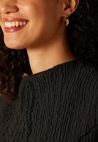 Black textured garment with a crinkled pattern, featuring a high neckline. Wearing small gold hoop earrings. Close-up of facial area.