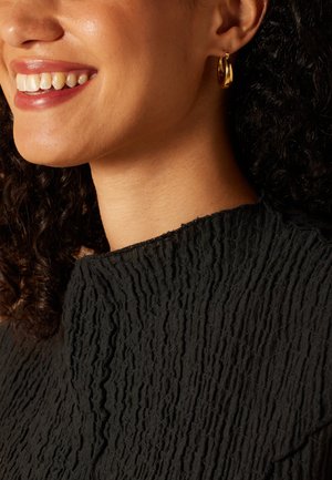 Black textured garment with a crinkled pattern, featuring a high neckline. Wearing small gold hoop earrings. Close-up of facial area.