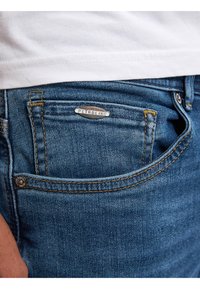 Jeans de mezclilla azul con costuras amarillas, que presentan una etiqueta de logo plateada y un pequeño bolsillo delantero. La tela parece texturizada y duradera.