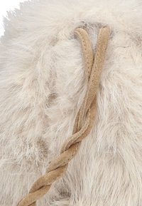 Cremefarbener Kunstpelz mit einer weichen, flauschigen Textur; verfügt über einen geflochtenen taupefarbenen Riemen zum Verschließen.