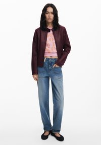 Burgundy leather jacket, pink graphic cropped shirt, loose blue jeans, black slip-on shoes with textured soles. Model stands against a white background.