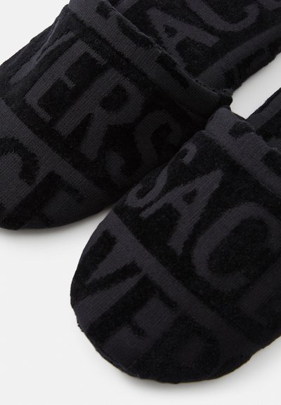 Μαύρες παντόφλες με πλούσια υφή και ανάγλυφο μοτίβο κειμένου, διαθέτοντας στρογγυλή μύτη και χαμηλό προφίλ. Το υλικό φαίνεται μαλακό και άνετο.