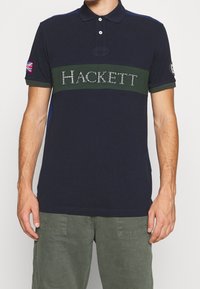 Mörkblå pikétröja i bomull med en grön rand som har "HACKETT" i vita bokstäver, korta ärmar och en britisk flagga-märke.