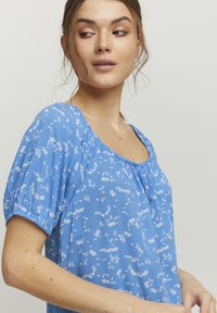 Blusa azzurro chiaro in tessuto morbido, con scollo quadrato, maniche corte a sbuffo e un motivo floreale bianco.