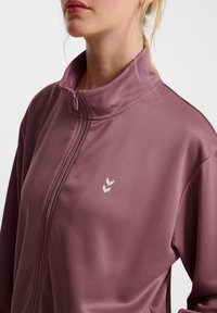 Lila zip-up jacka med hög krage, slät textur och vit logotypdetalj på bröstet. Har en sportig, figursydd design.