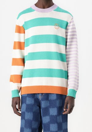 Homme portant un pull rayé multicolore avec des rayures bleu sarcelle, blanc, orange et lavande, assorti à un jean à carreaux bleu.
