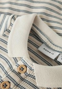Polo in cotone beige con strisce blu, dotato di colletto a coste e due bottoni marroni al collo. Etichetta del marchio visibile all'interno.