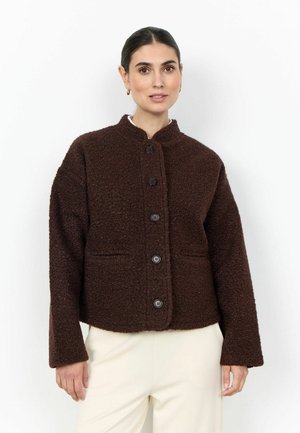 Vrouw draagt een donkerbruine wollen jas met knopen en lange mouwen en lichtbeige broek, staand tegen een witte achtergrond.