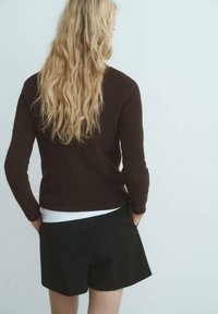 Pull marron à côtes avec des manches longues, associé à un short noir ajusté. La tenue comprend un haut blanc superposé sous le pull.