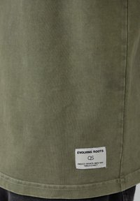 Oliwkowa bawełniana koszulka z krótkim rękawem, prostym dołem i metką z napisem "EVOLVING ROOTS" wszytą blisko dolnej krawędzi.