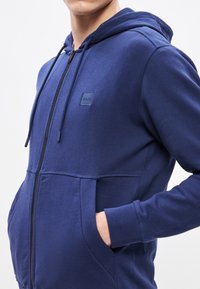 Tengerészkék, cipzáras kapucnis pulóver puha anyagból, elöl zsebbel, húzózsinóros kapucnival és egy kis logópáttal a mellkason.