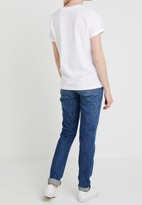 Person, der står mod en hvid baggrund iført en simpel hvid t-shirt, blå oprullede jeans og hvide sneakers, set fra bagved.