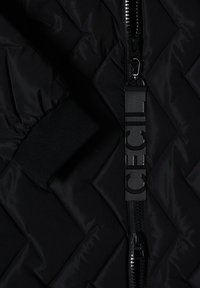 Section de veste matelassée noire avec fermeture éclair visible et languette en tissu noir comportant des lettres embossées partiellement visibles.