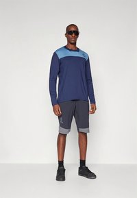 Tengerészkék hosszú ujjú ing világoskék díszítéssel és logóval; szürke rövidnadrág mintás szegéllyel; fekete sportcipő; sötét napszemüveg.