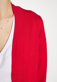 Cárdigan de punto rojo con cuello en V, textura fina, diseño abierto en la parte delantera y acentos acanalados en los bordes. Capa interior blanca visible en el cuello.